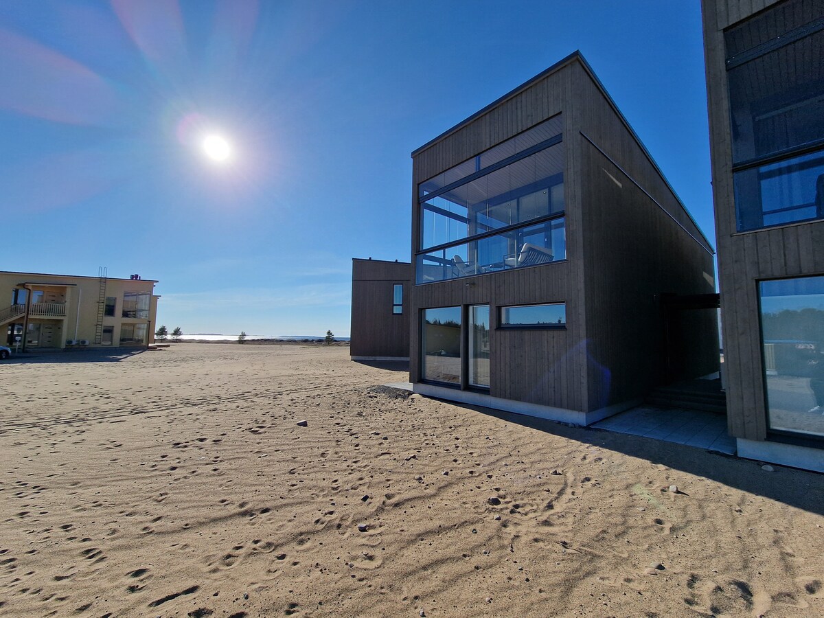 The exterior of the modern two-story building is framed by a sandy landscape under a clear blue sky. Large glass windows reflect sunlight, creating a bright and airy appearance. The structure's wooden facade complements the natural surroundings.