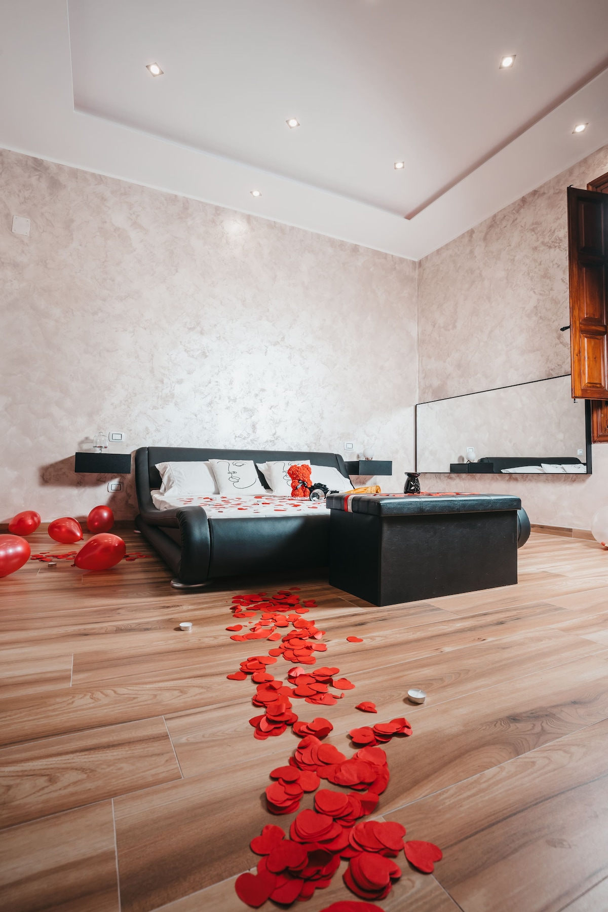A stylish room features a black sofa adorned with decorative pillows. A path of red rose petals leads to a coffee table, surrounded by scattered balloons. Natural light enters through a window with a wooden frame, illuminating the light-colored walls and warm wooden flooring.