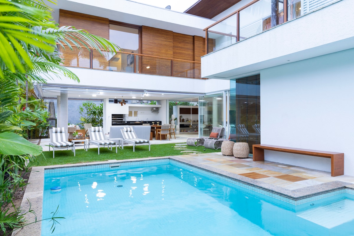 The image features a modern pool area surrounded by lush greenery. A clear blue pool reflects the sky, with several lounge chairs positioned nearby. The adjacent outdoor dining space is visible through glass doors, leading to a well-equipped kitchen and living area.