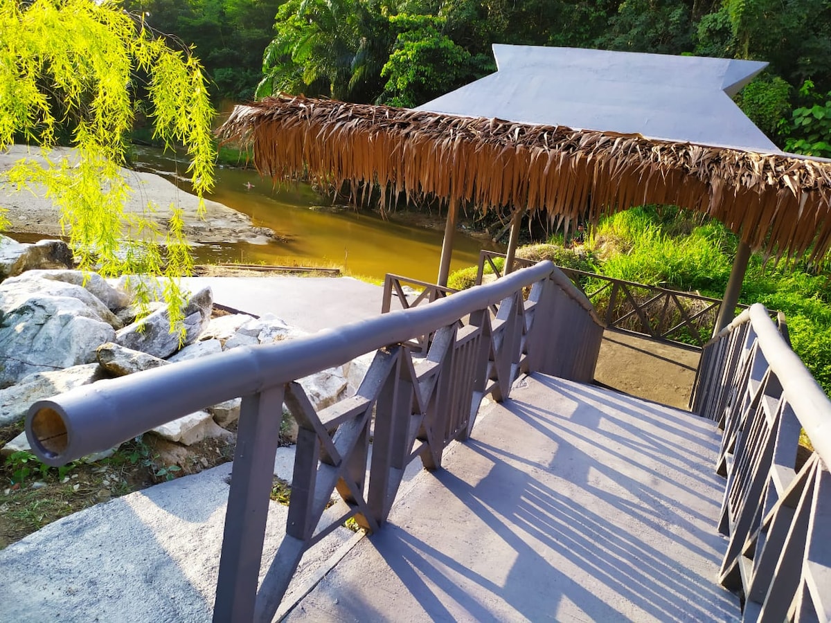 A rustic wooden pavilion with a thatched roof overlooks a tranquil river. A sloped wooden walkway leads down from the pavilion, surrounded by lush greenery and large rocks, inviting relaxation in nature.