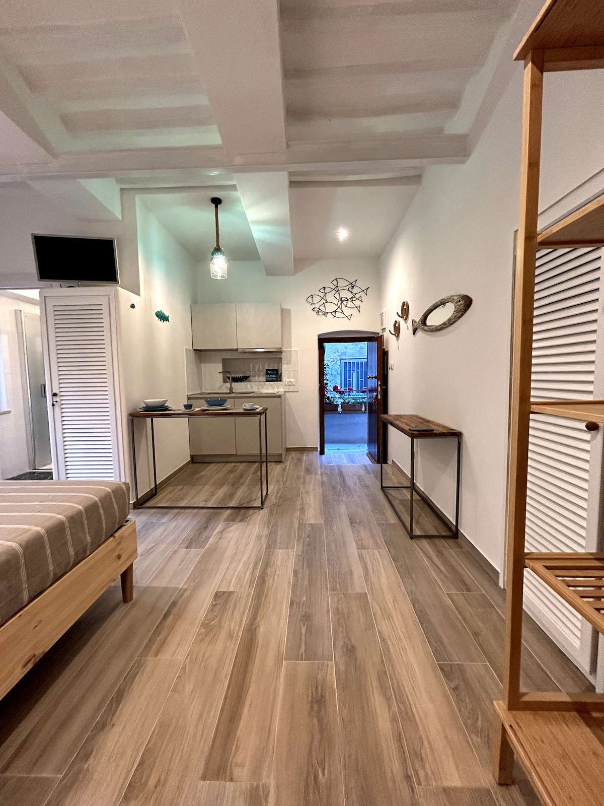 A modern studio space is presented with light wooden flooring and a comfortable bed. A minimalist kitchenette is featured with sleek cabinetry. Decorative mirrors and wall art contribute to the ambiance, while a doorway at the far end leads to a charming exterior view.