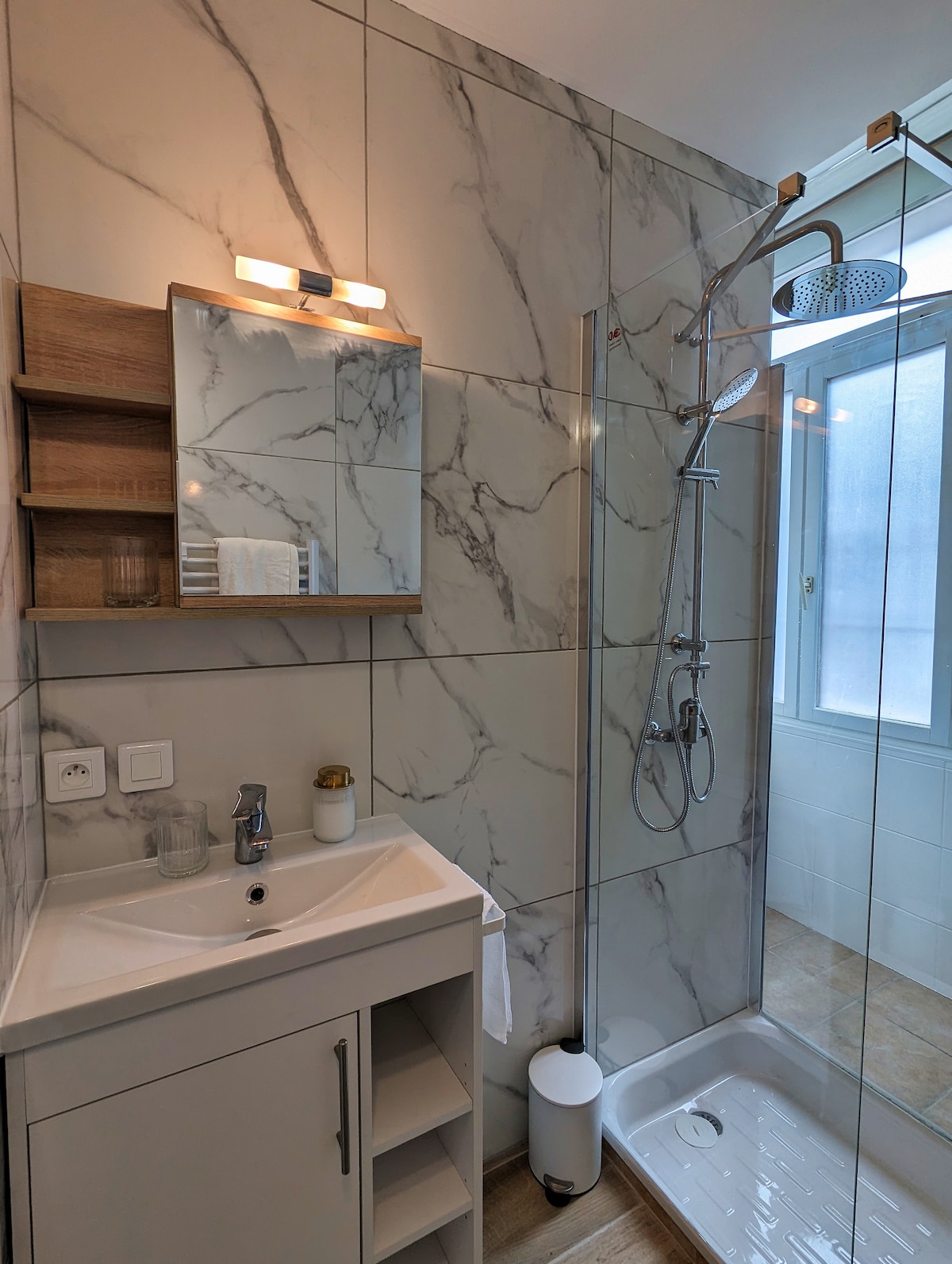 A modern bathroom features sleek marble-patterned walls and a glass shower enclosure. A wooden shelf holds neatly arranged toiletries. A small sink with a contemporary faucet and storage underneath is positioned beside a well-lit mirror, reflecting natural light from a nearby window.