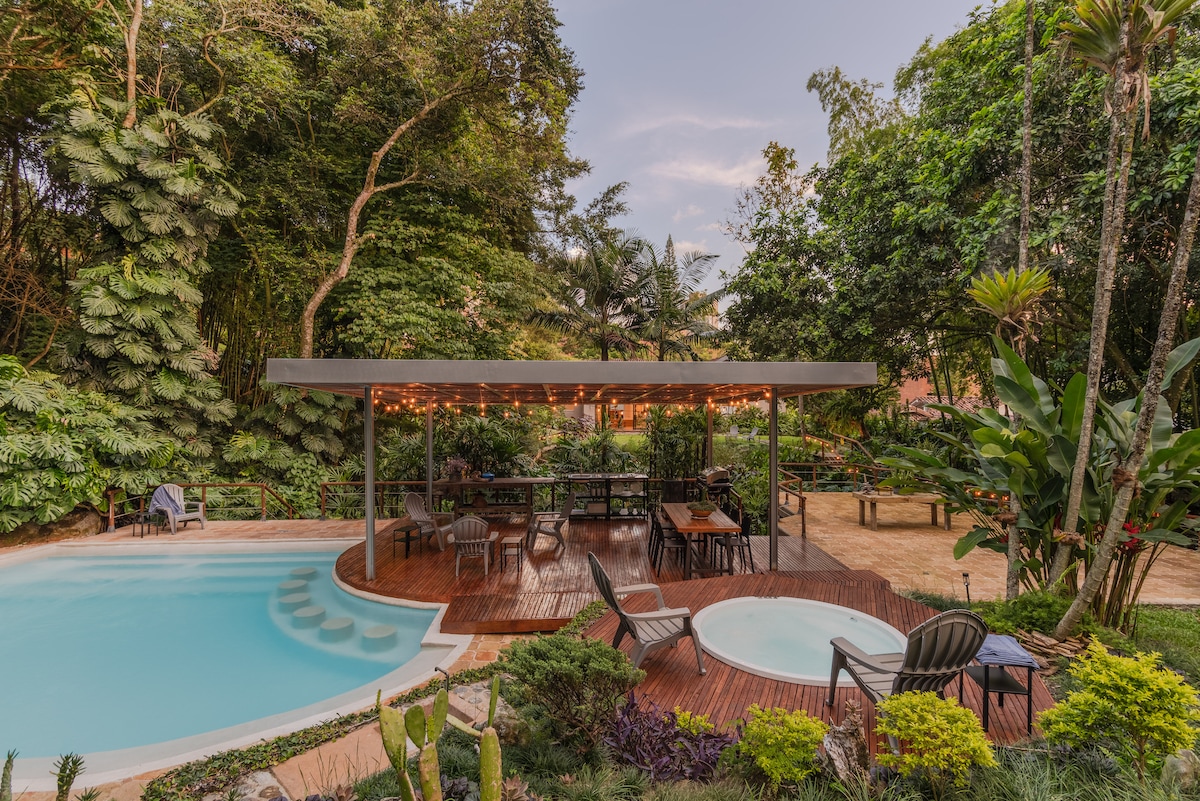 A spacious pool area is featured with a tranquil swimming pool and a jacuzzi, surrounded by lush greenery. A shaded wooden deck is furnished with multiple tables and chairs, offering a relaxing space for outdoor dining. Soft lighting enhances the gentle ambiance of the garden.