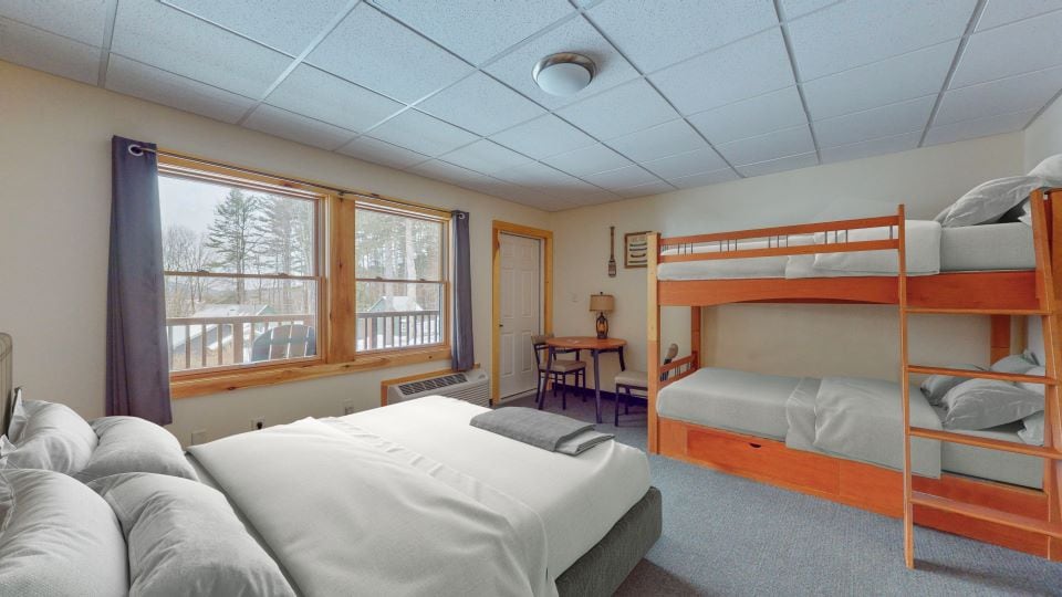 A spacious room features a queen bed and twin bunk beds, ideal for family accommodations. Large windows provide natural light and a view of the outdoors. A small table and chair are positioned near the window, enhancing the functional layout.