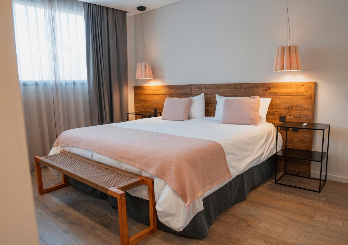 A spacious bedroom features a wooden headboard and two pendant lights on either side. A light pink throw blanket drapes over the bed. A wooden bench is positioned at the foot of the bed, and sheer curtains allow soft light to filter in.