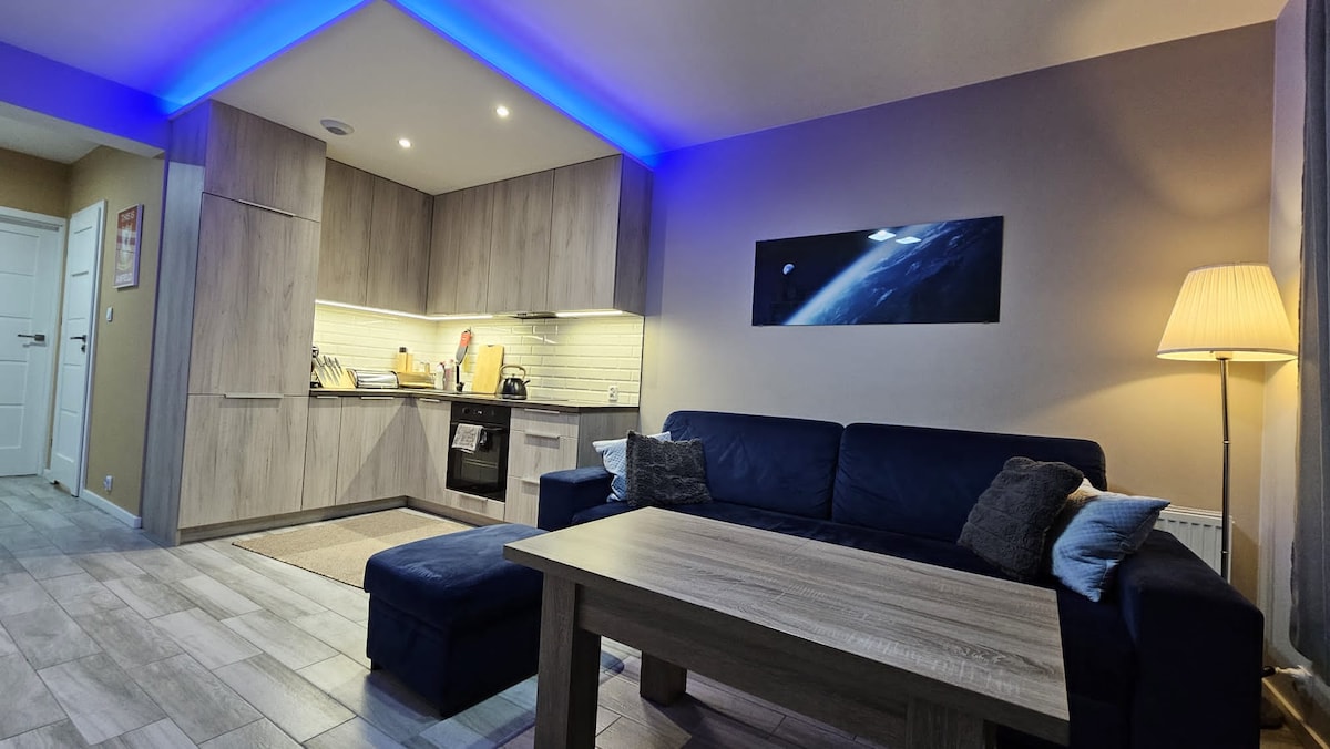 A modern living area features a cozy navy sofa paired with a wooden coffee table. A softly lit kitchen is visible in the background, showcasing sleek cabinetry and essential appliances. Soft blue lighting enhances the ambiance, while a warm floor lamp adds additional illumination.