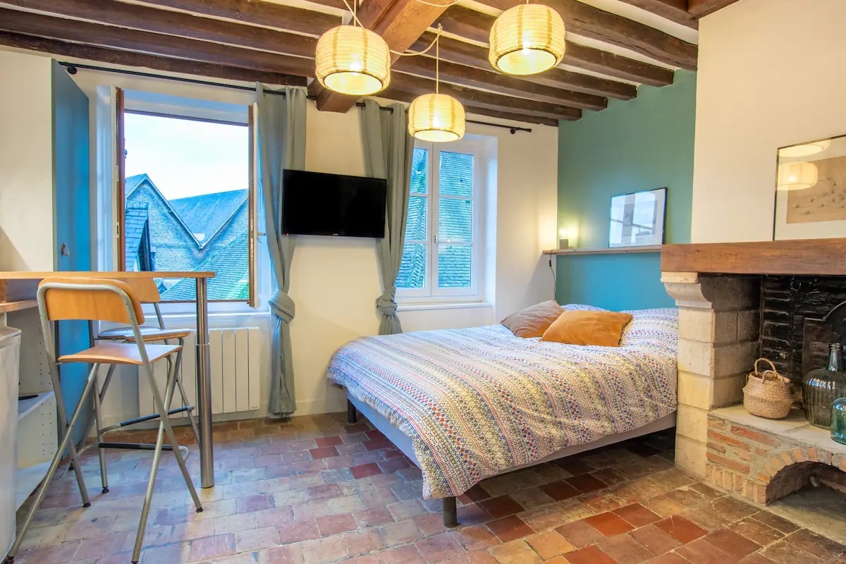A cozy bedroom features a spacious bed covered in a patterned blanket, complemented by soft lighting from pendant fixtures. A window provides natural light, with a view of the rooftops outside. A small dining area and a decorative fireplace enhance the inviting atmosphere.