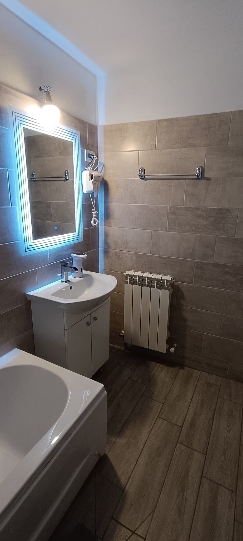 The bathroom features modern, neutral-toned tiles. A large mirror with integrated lighting is mounted above a sleek vanity with a sink. A bathtub is included, and a towel rack is positioned on the wall, with a radiator nearby for warmth.