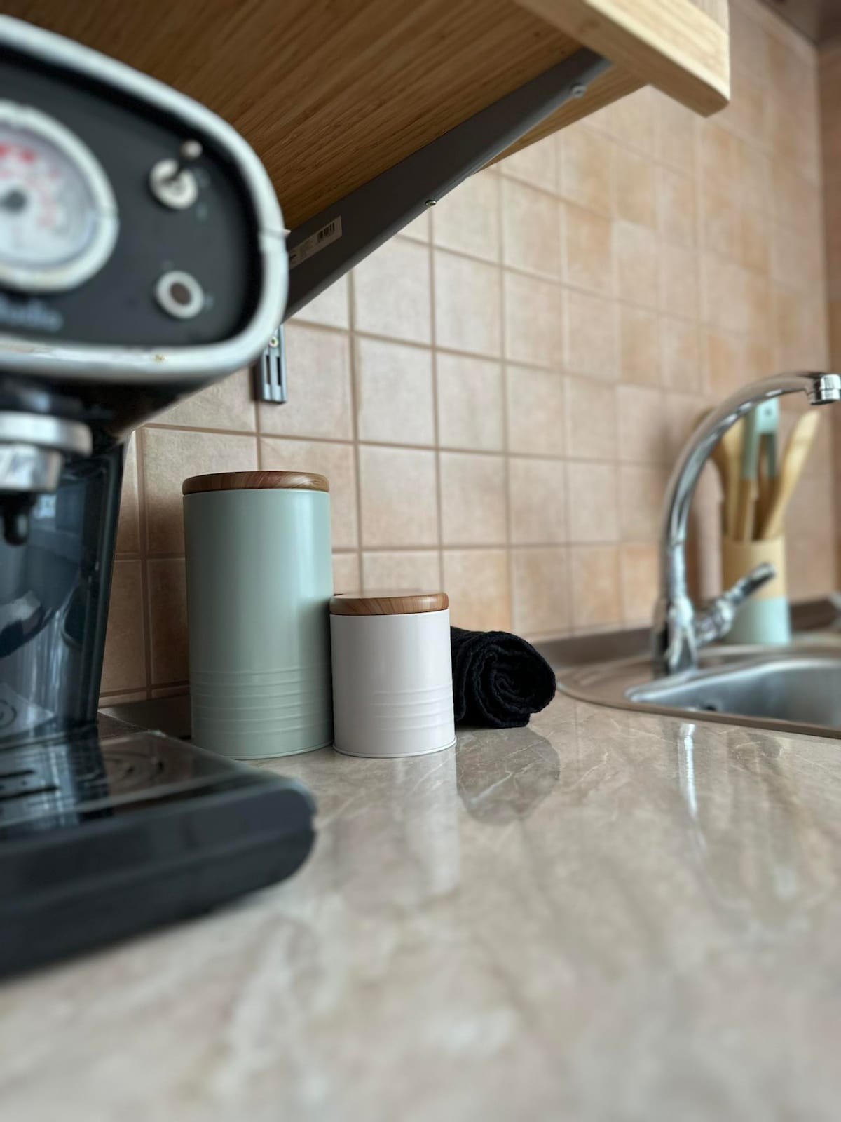 A modern kitchen countertop is displayed, featuring a coffee machine alongside two cylindrical canisters with wooden lids. A black towel is neatly placed near a stainless steel sink, which is surrounded by utensils in a wooden holder. The backsplash consists of light-colored tiles.
