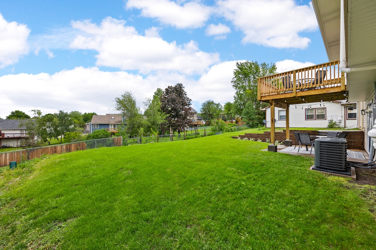 A spacious backyard is shown, featuring a large green lawn and a deck with outdoor seating. Nearby properties can be seen in the background, along with mature trees and landscaping that enhance the serene outdoor environment.