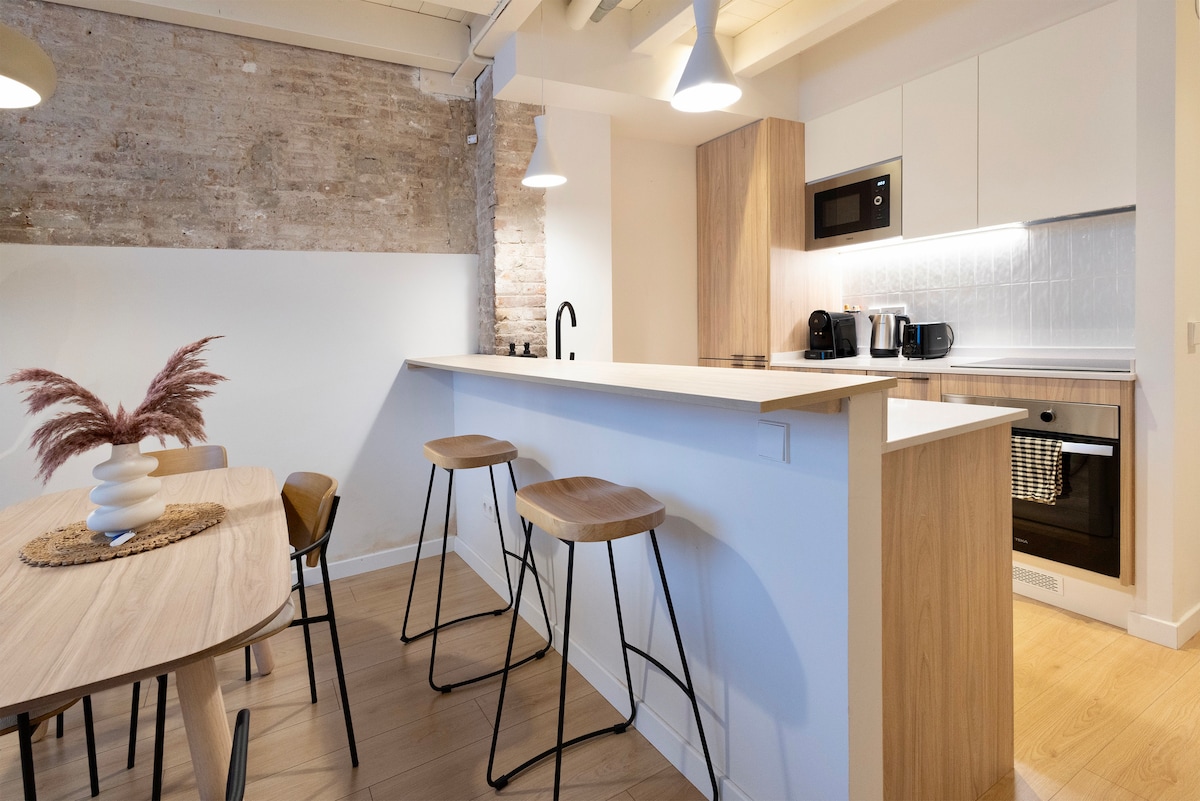 A modern kitchen area is highlighted with wooden cabinetry and white countertops. Three bar stools are positioned at a sleek counter, enhancing the dining space. Natural light reflects off the light wood flooring, creating an open and airy ambiance.