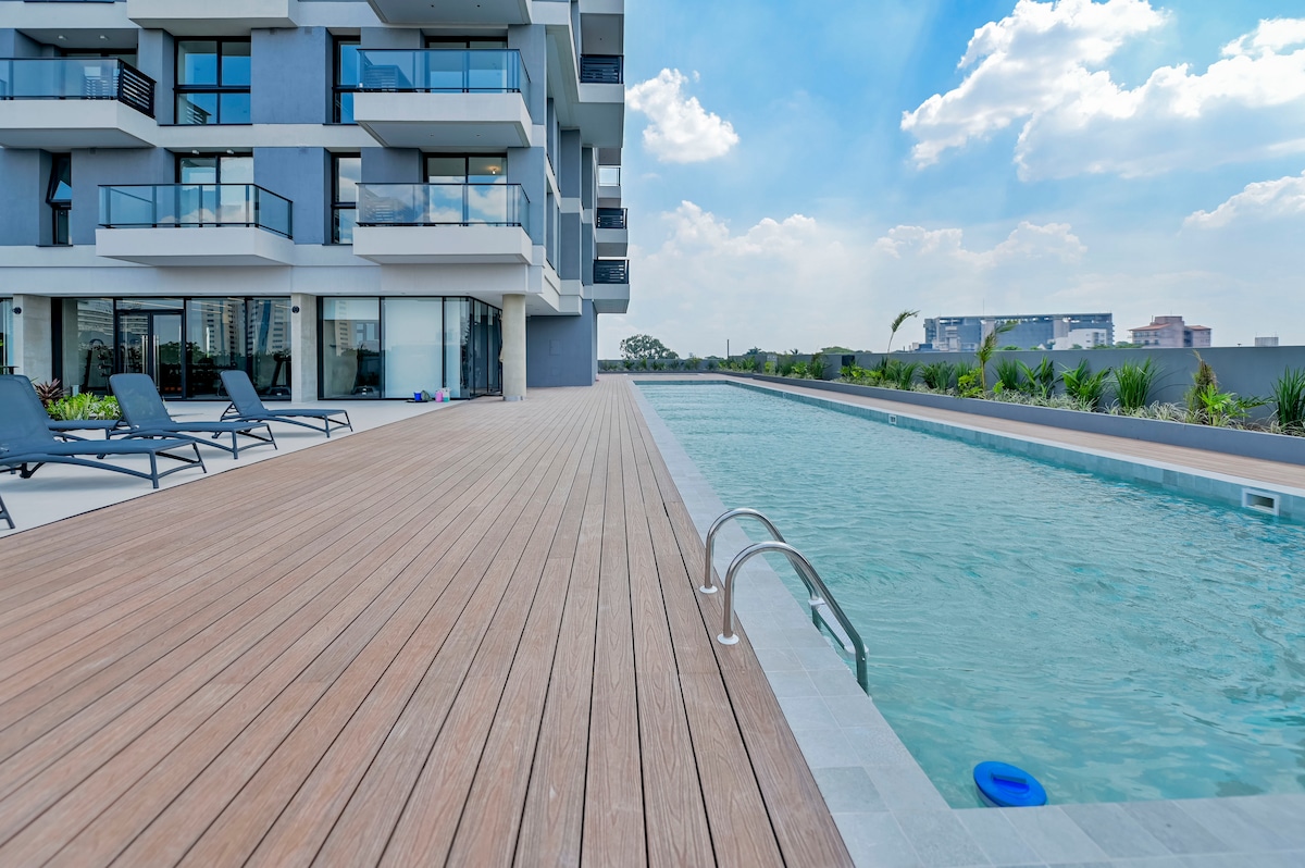 A semi-Olympic swimming pool is visible, bordered by a wooden deck and lined with poolside lounge chairs. The modern building features multiple balconies overlooking the pool area, with lush greenery placed along the perimeter enhancing the surrounding ambiance.