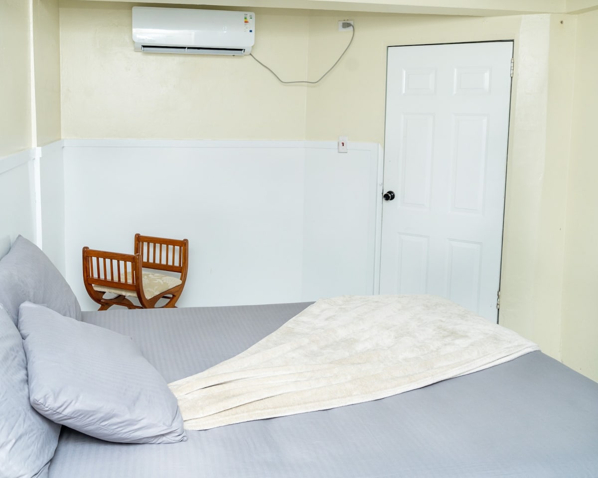 A spacious bed is featured with soft gray bedding, complemented by a neatly arranged light-colored throw. A small wooden chair and table occupy a corner, with a white door visible in the background. An air conditioning unit is mounted on the wall.