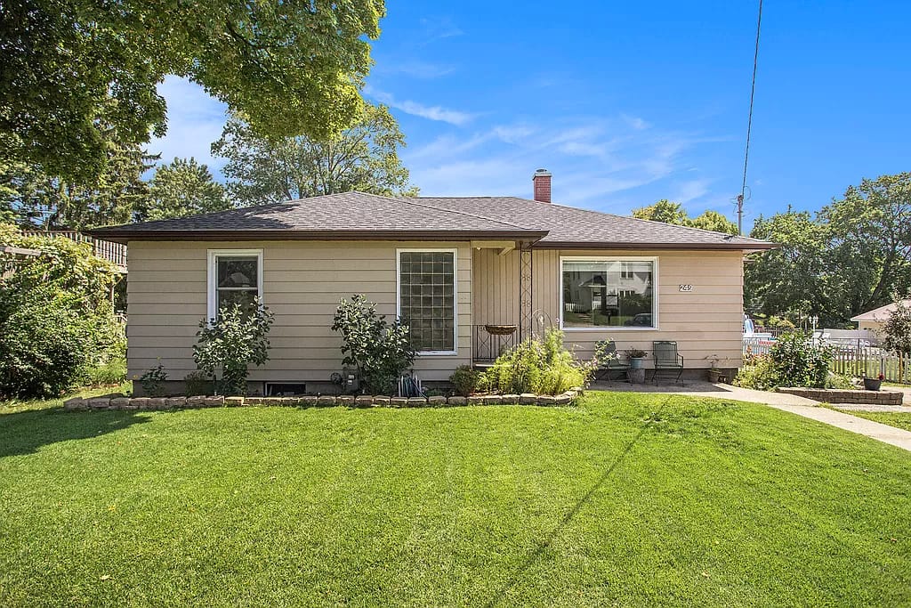 A single-story house features a neat exterior with light-colored siding and a well-maintained lawn. Green shrubs and small plants frame the entrance, while a few outdoor chairs are positioned on the porch, offering a welcoming outdoor space.