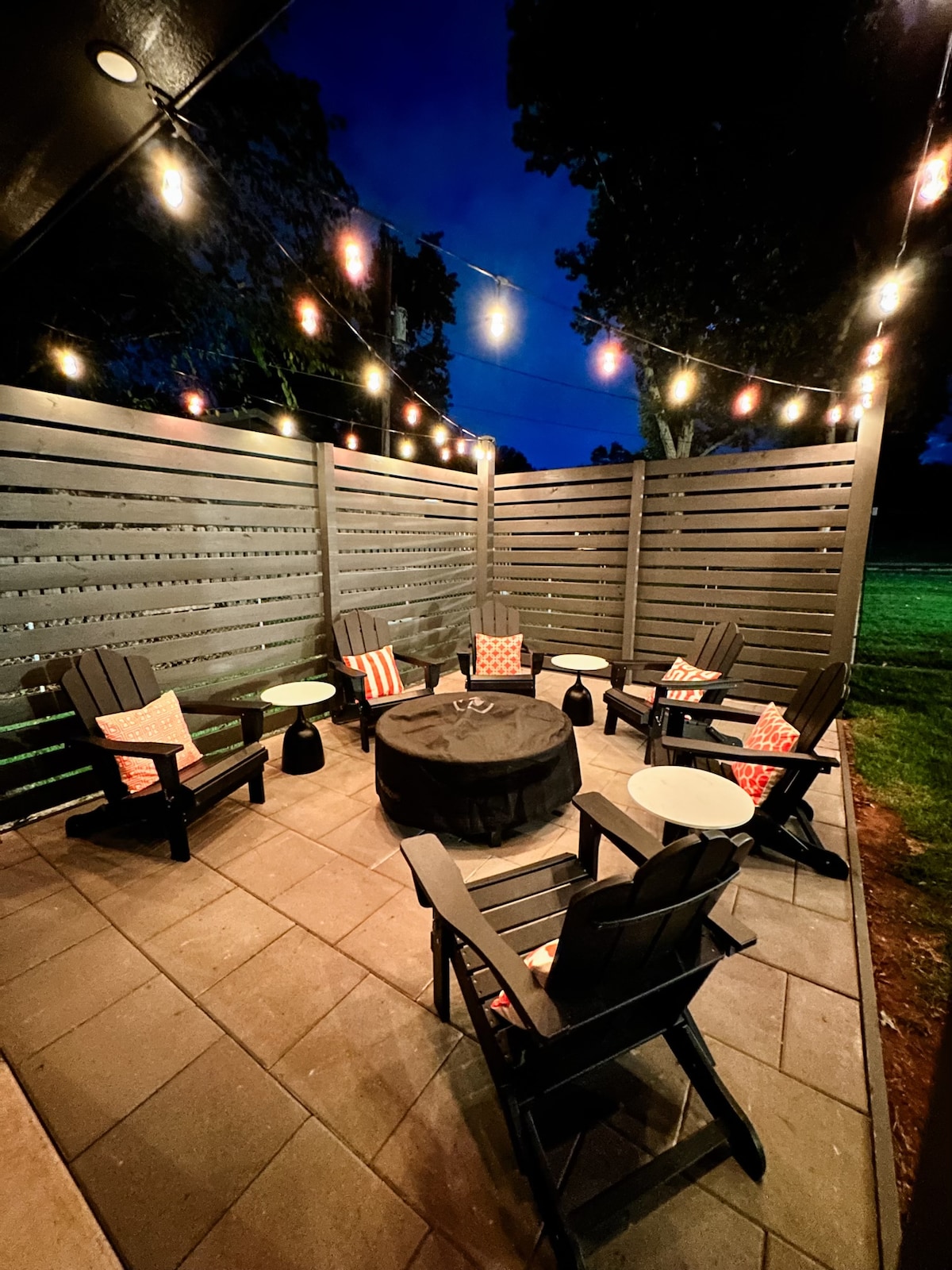An outdoor gathering space is enclosed with wooden slats, featuring a fire pit at the center. Surrounding the fire pit are six comfortable chairs with cushions. String lights hang overhead, creating a warm ambiance in the evening, while the lawn is visible beyond the enclosure.