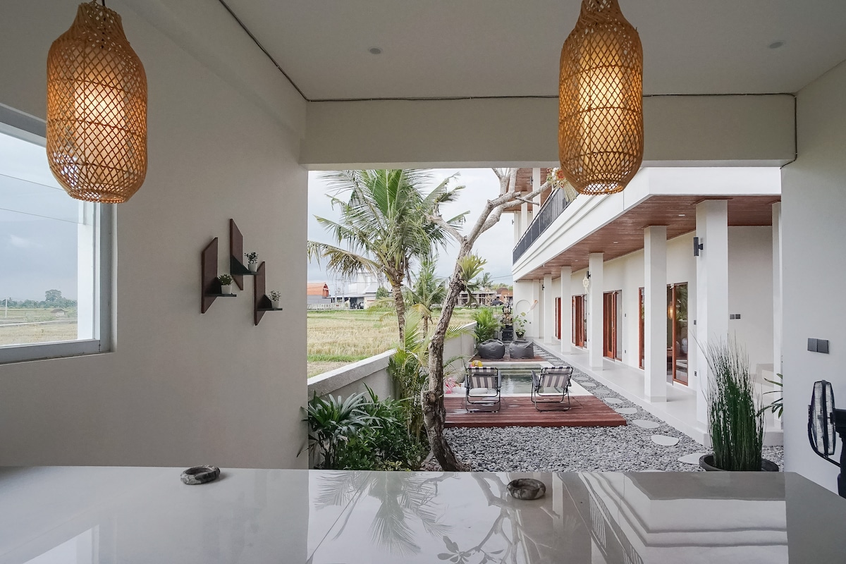 A view from inside the villa showcases an open space leading to an outdoor area. Natural light fills the room, where crafted light fixtures hang elegantly. A spacious patio with greenery extends towards a pool, framed by palm trees and vibrant vegetation.