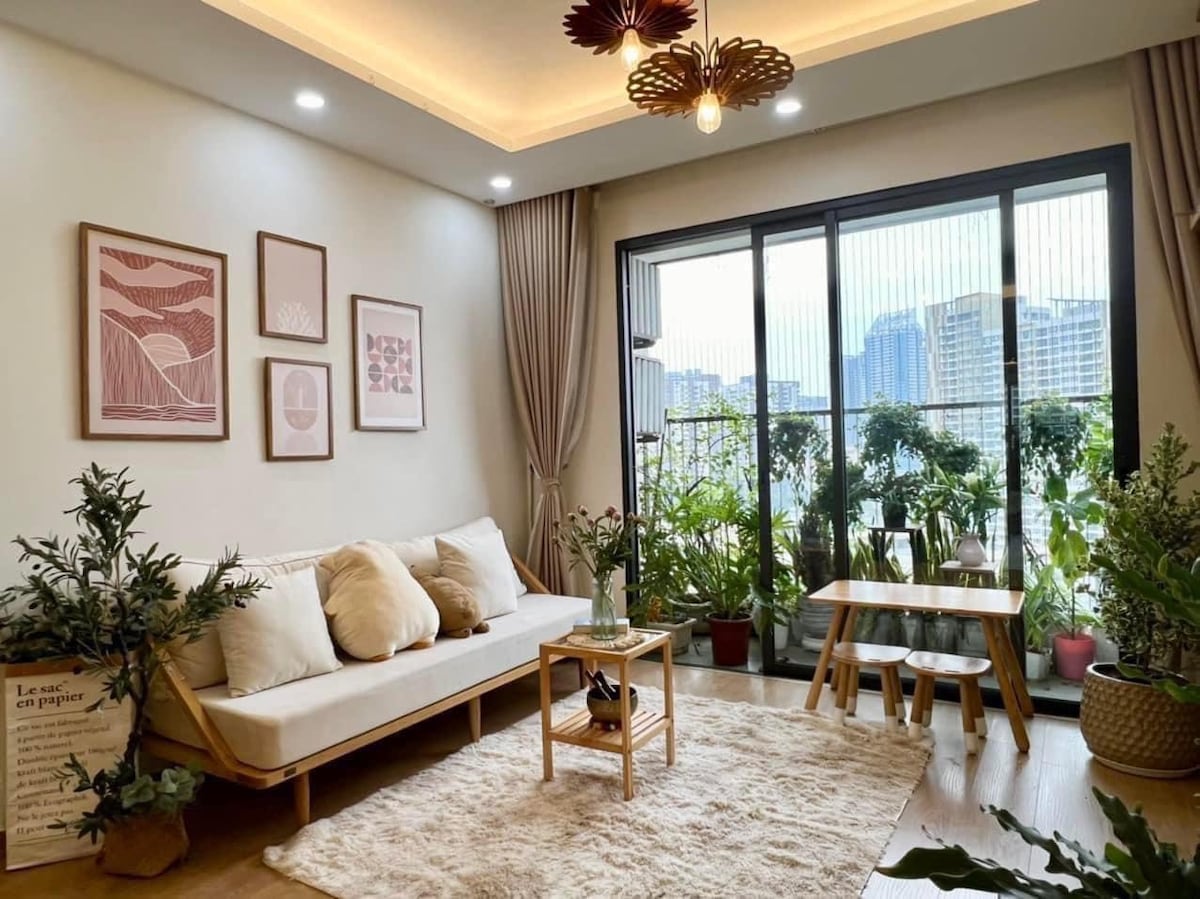 A light-filled living area features a modern sofa with decorative cushions, positioned next to a small wooden table. Indoor plants are situated around the space, enhancing the connection to nature. Floor-to-ceiling glass doors open to a balcony with views of the city skyline.