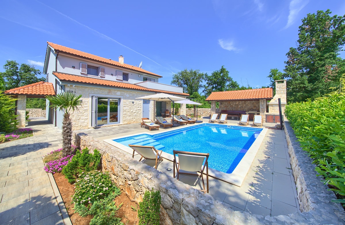 An inviting exterior view of the villa featuring a spacious private pool surrounded by lounge chairs. Lush greenery and colorful flowers enhance the area, while a covered outdoor dining space is visible alongside the house. The structure’s two stories are accentuated by a red-tiled roof.