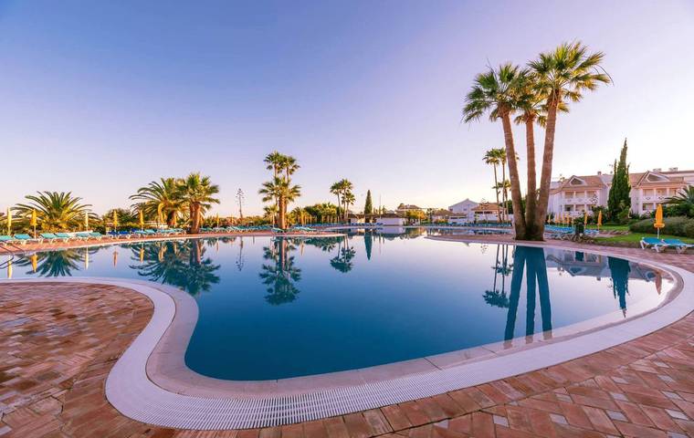 A tranquil swimming pool reflects the surrounding palm trees and clear sky. Lounge chairs are positioned along the water's edge, inviting relaxation in a serene environment. The clean, curved pool design enhances the spaciousness of the outdoor area.
