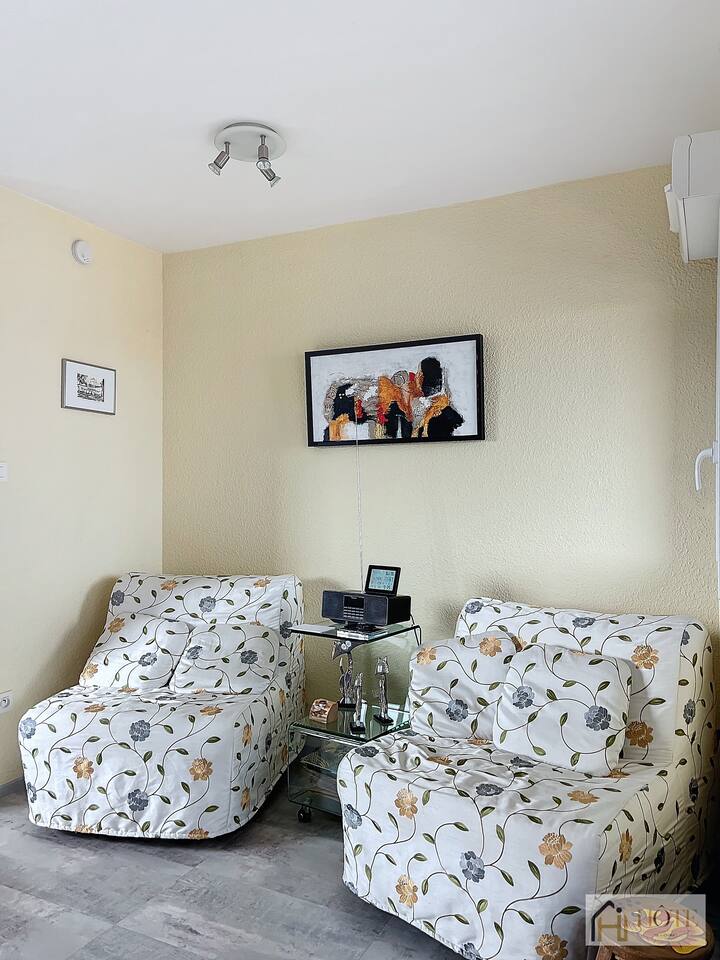 нека биде 150 знака


Влезете во светол простор за живеење со две цветни фотелји, стаклена маса со технологија и шарена уметност. Топлите жолти ѕидови и сивите акценти создаваат удобно место за опуштање.