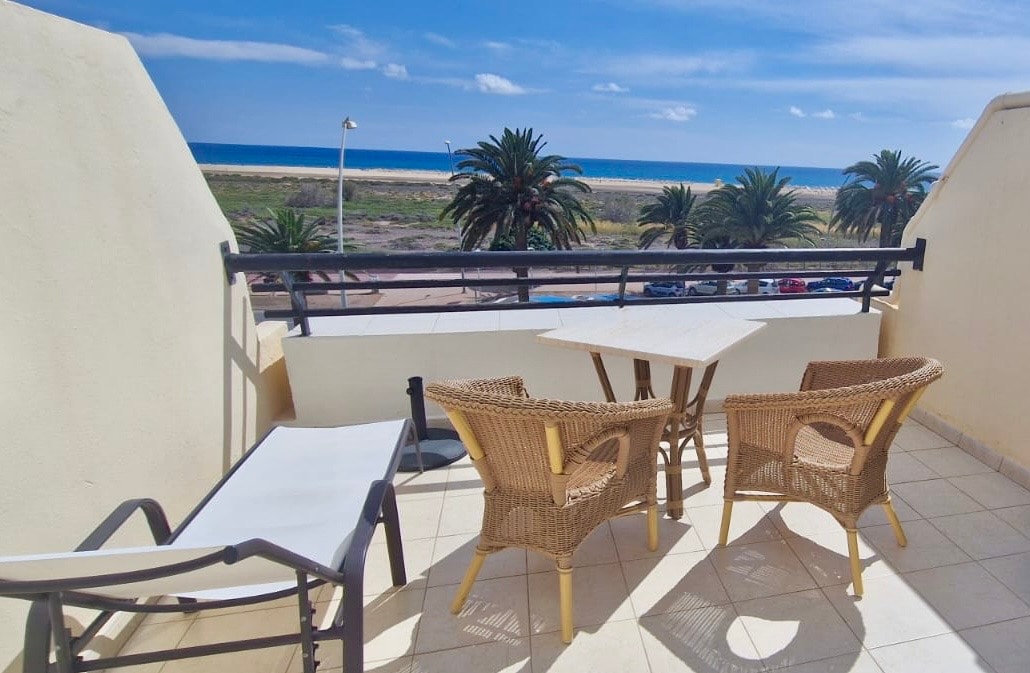 A terrace is visible, featuring a small table and two wicker chairs, accompanied by a chaise lounge. Beyond, the Atlantic Ocean can be seen, along with palm trees lining the shore. The clear blue sky enhances the coastal view.