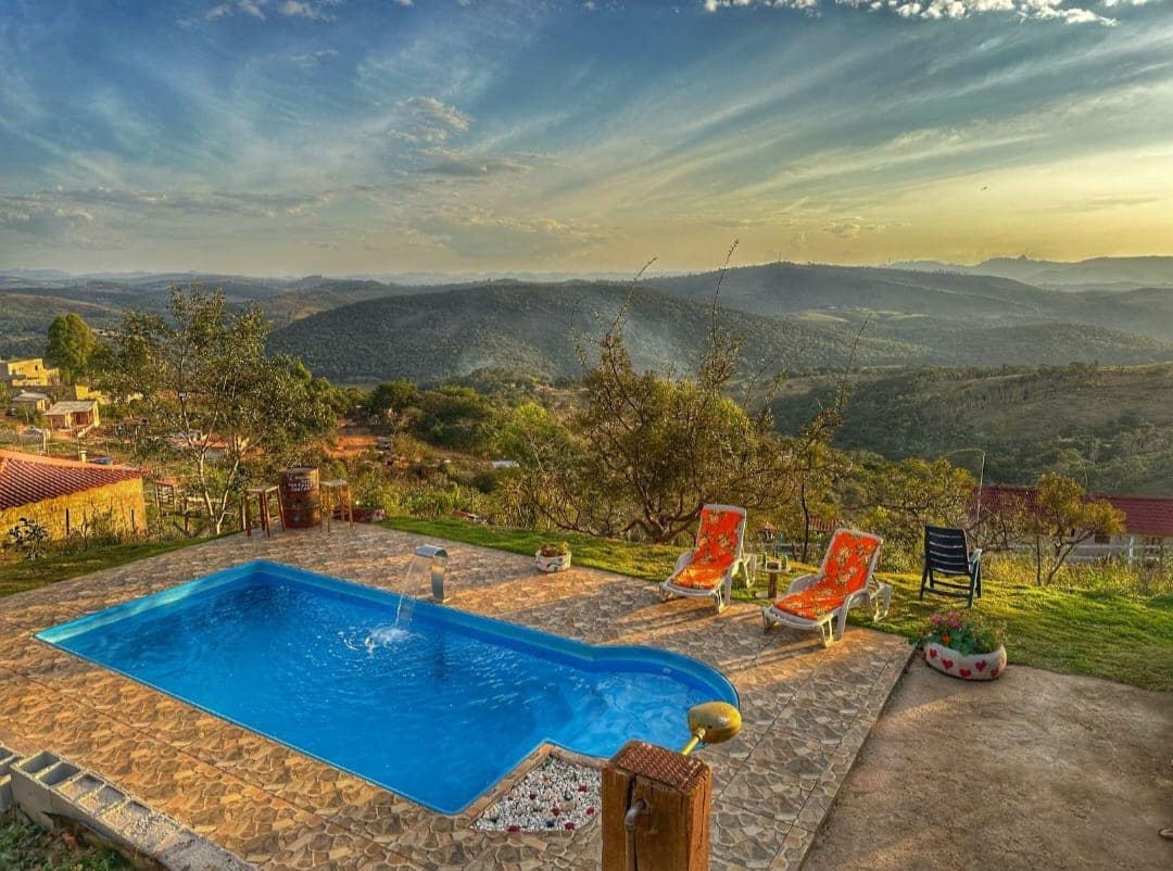 A swimming pool is situated on a terrace overlooking sweeping mountain views. Two lounge chairs are positioned beside the pool, which is framed by lush greenery. The sky features soft hues of orange and blue, indicating a serene sunset.