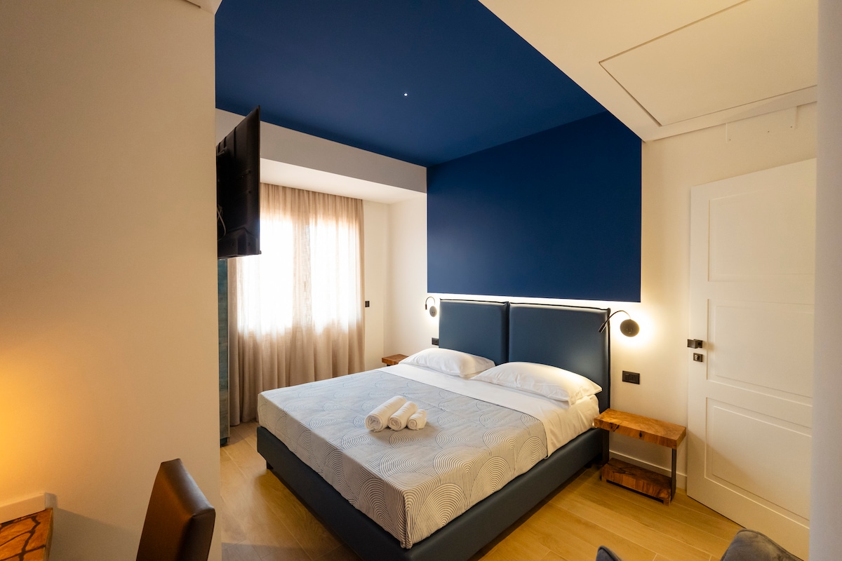 A welcoming bedroom features a spacious bed with crisp white linens, framed by a deep blue accent wall. Natural light filters in through a window dressed with sheer curtains, and a television is mounted on the opposite wall. Two wooden bedside tables sit beside the bed.