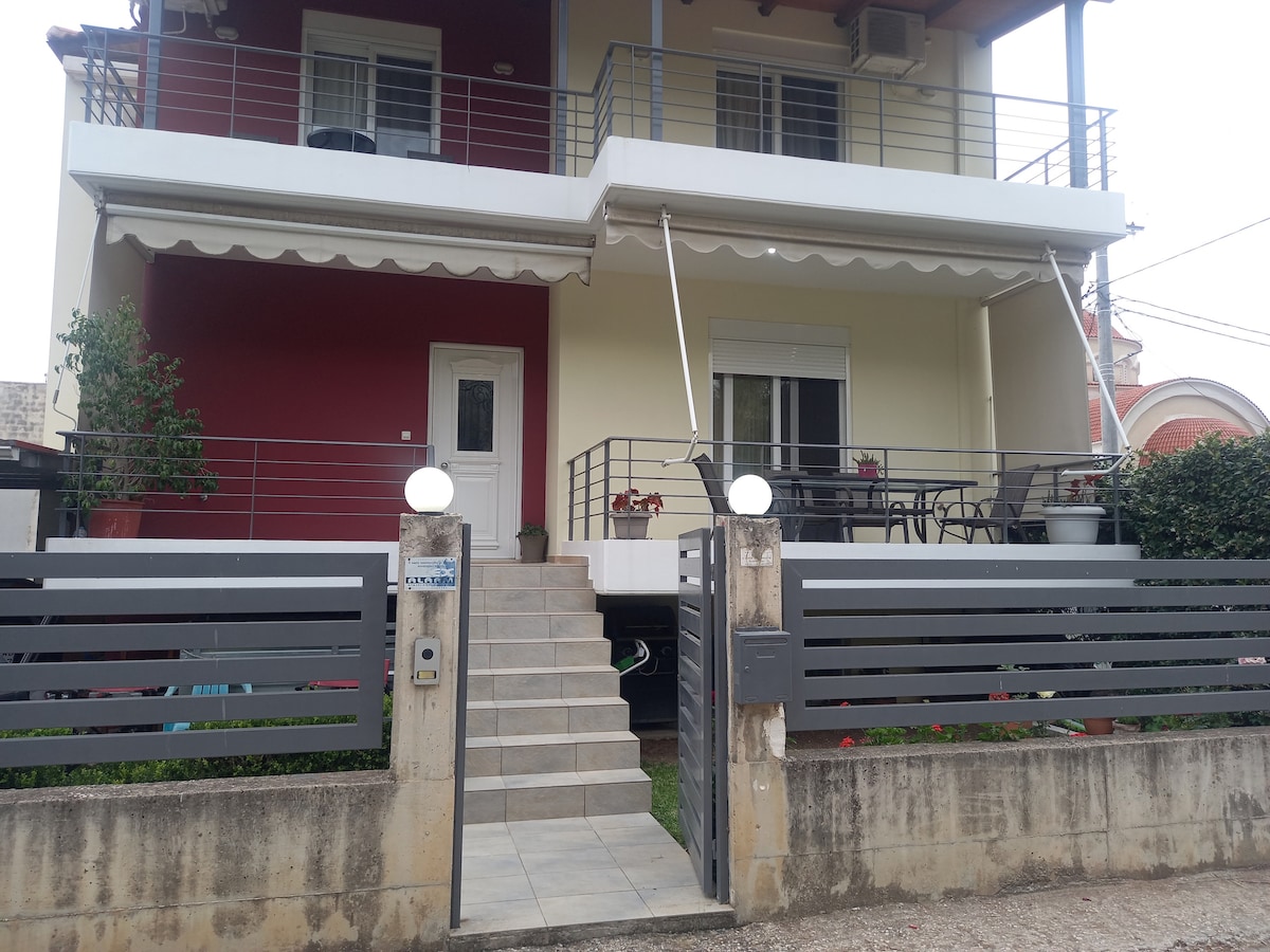 A multi-story building is displayed, featuring a modern facade with a combination of red and cream colors. A set of stairs leads to the entrance, which is flanked by planters. Outdoor seating is visible on the balcony, along with railings that provide a spacious look.