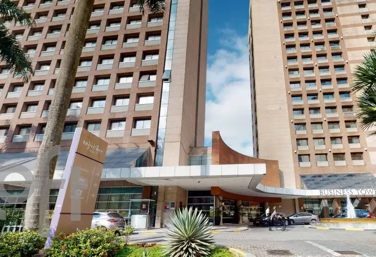 The exterior of a modern hotel building is shown, featuring a welcoming entrance and large glass windows. The design includes a blend of concrete and wood elements, surrounded by palm trees and greenery, with vehicles parked nearby.