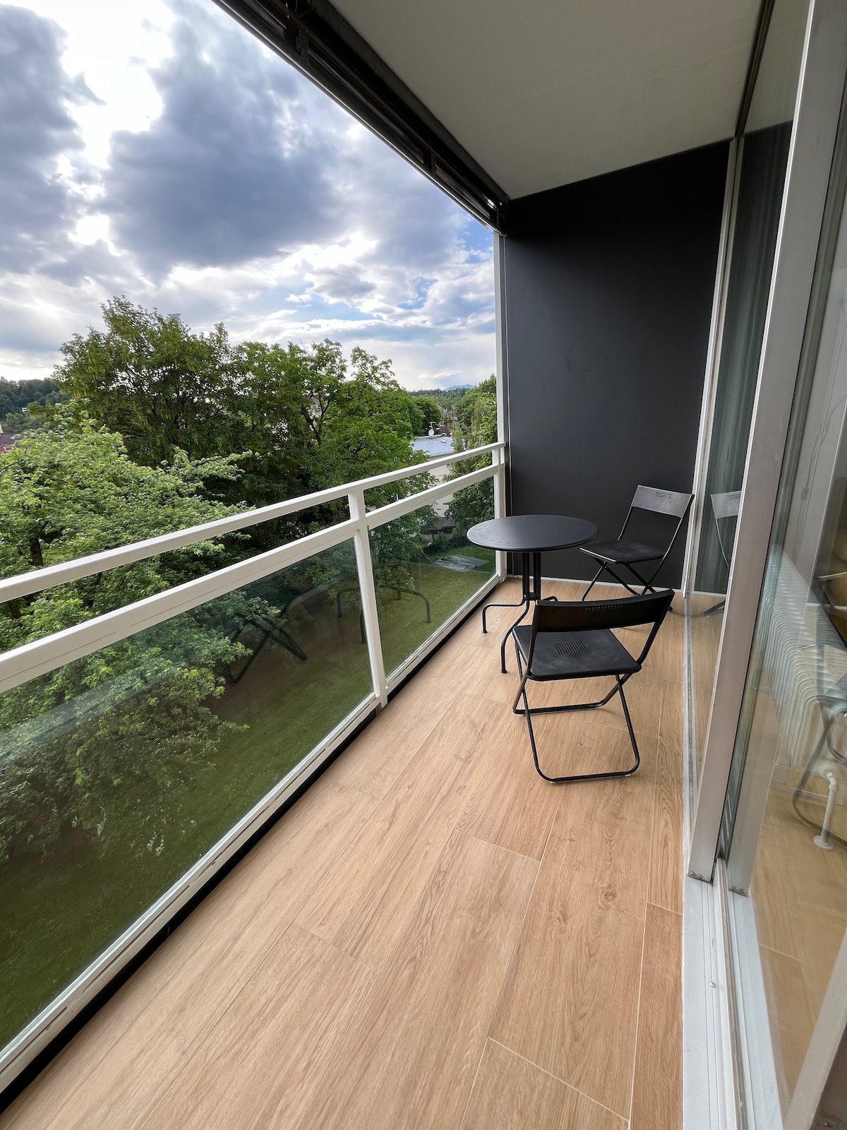 A west-facing balcony features a small round table and two black chairs, complemented by a serene view of greenery. The glass railing allows abundant natural light while offering privacy from neighboring spaces. The wooden flooring adds warmth to the outdoor area.