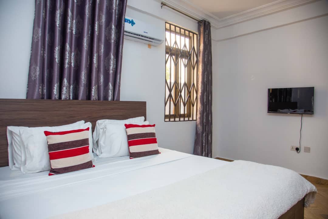 A well-lit sleeping area features a spacious bed with white linens and decorative pillows in red and grey stripes. Light streams in through a window adorned with elegant curtains. A wall-mounted TV is positioned across from the bed, enhancing the room's functionality.