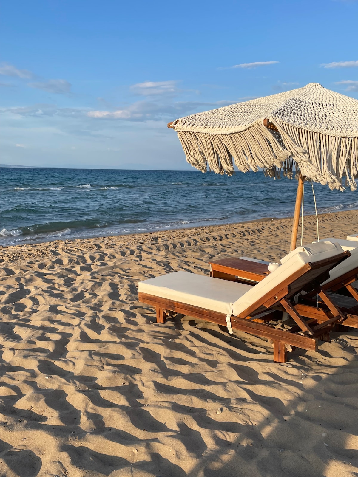 Two wooden lounge chairs are positioned on a sandy beach, accompanied by a large umbrella with fringe detailing. The ocean is visible in the background, with gentle waves lapping at the shore and a clear blue sky above.