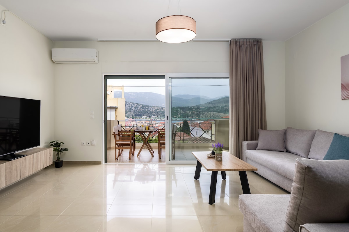A spacious living area is furnished with a comfortable sofa and a wooden coffee table. Large glass doors open to a balcony, presenting scenic views of the surrounding landscape. Natural light fills the room, while soft curtains add a touch of elegance. An air conditioning unit is mounted above.