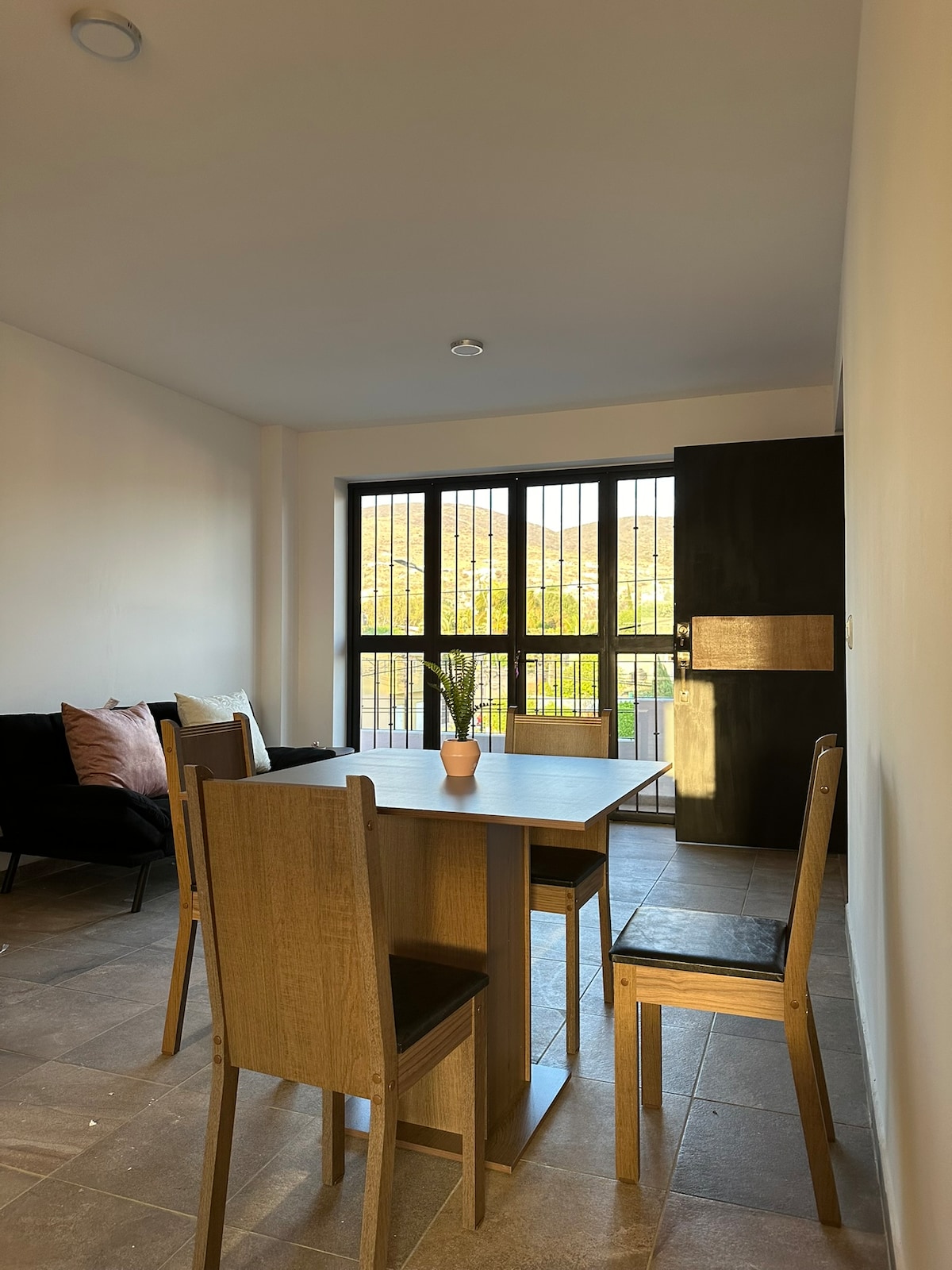 A dining area features a wooden table surrounded by four chairs. Large windows allow natural light to fill the room, with a view of greenery visible outside. A small potted plant sits on the table, adding a touch of nature to the space.