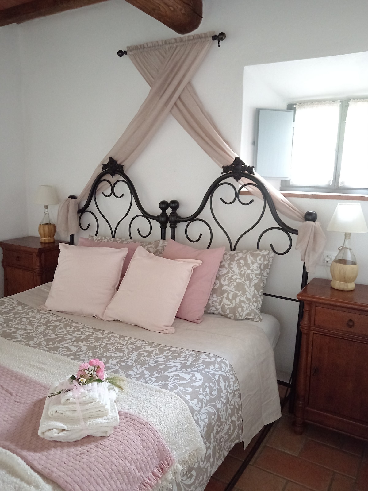 A spacious bed is framed by an elegant black iron headboard adorned with soft drapes. Light pink and cream decorative pillows complement the neutral-colored linens. Two wooden nightstands with lamps are positioned on either side, while a window offers natural light to the inviting room.