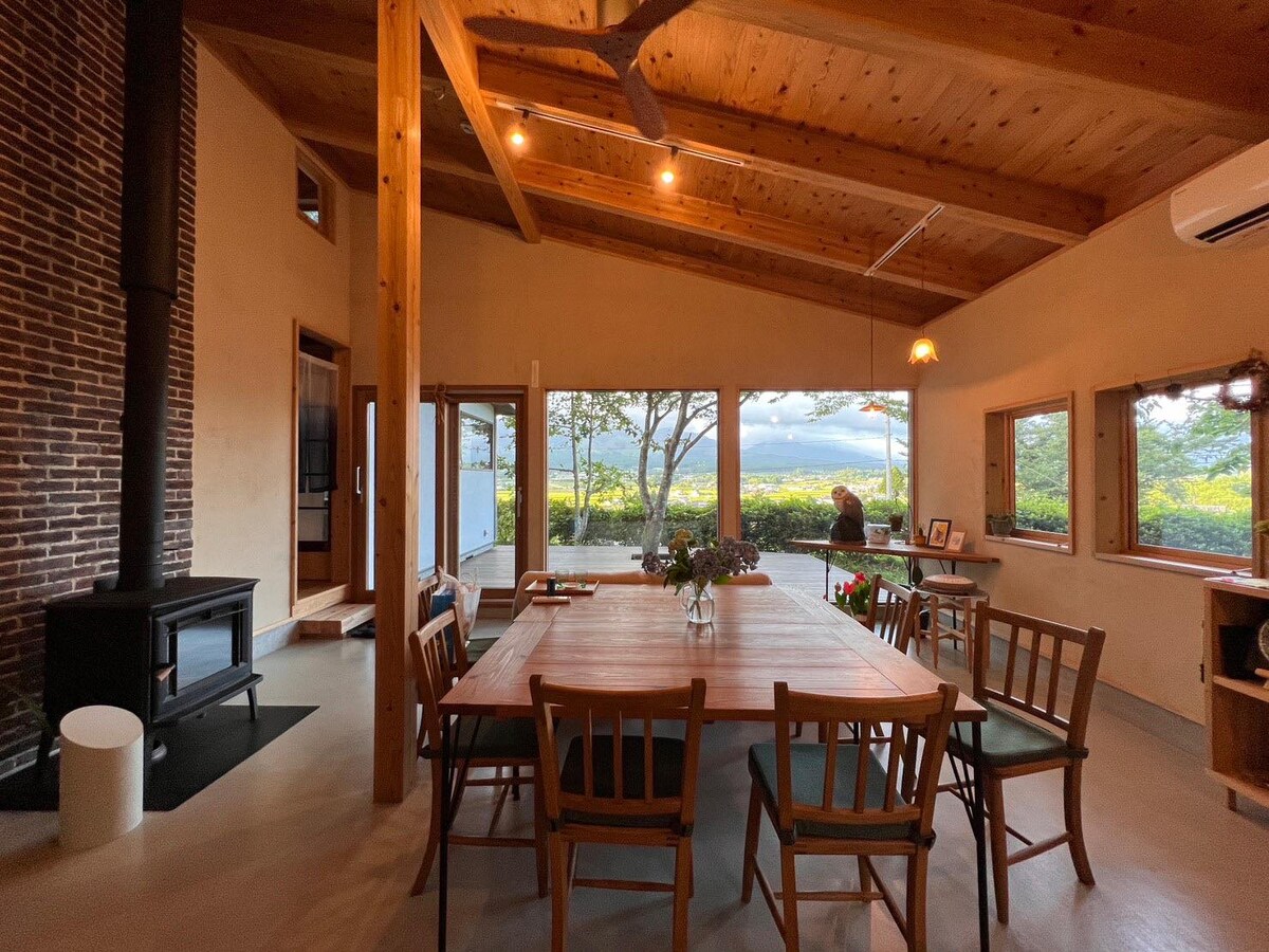 A spacious dining area features a large wooden table surrounded by several chairs, with a view of the landscape visible through large windows. A wood-burning stove is positioned next to a brick wall, enhancing the room's natural elements and warmth.