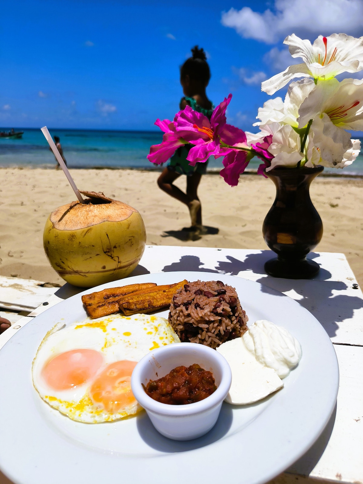 A plate of breakfast featuring two fried eggs, black beans, rice, and a dollop of sour cream is presented on a white table. A coconut with a straw and a small vase of pink flowers complement the scene, with a beach and sea visible in the background.