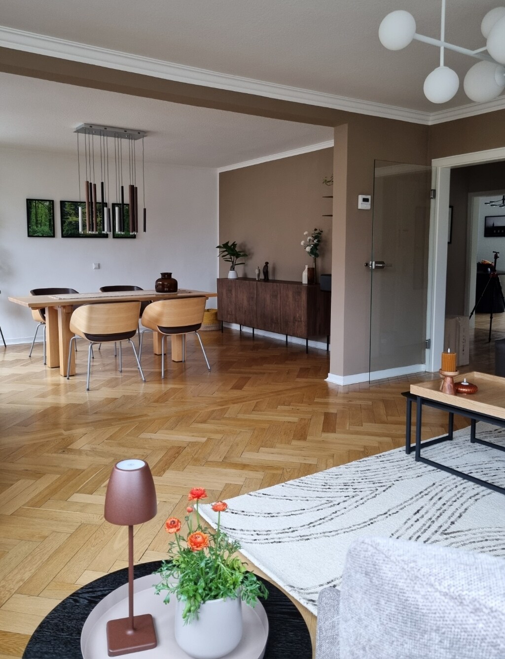 A spacious living and dining area is presented, featuring a rectangular dining table surrounded by four wooden chairs. A mid-century style lamp and potted plants are placed on a low coffee table. Light-colored walls contrast with the rich wood flooring, enhancing the room's warmth.