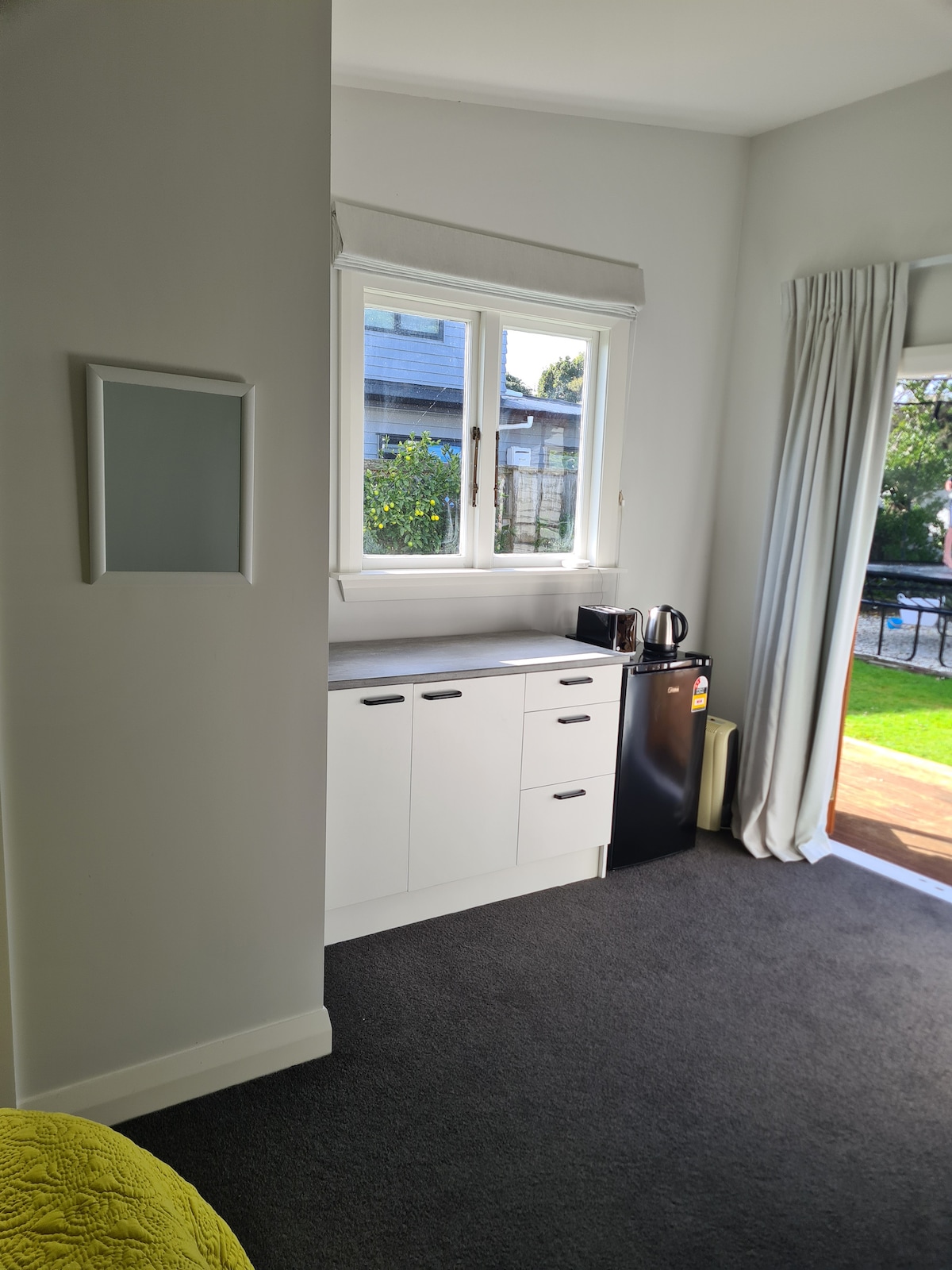 A kitchenette is seen with a white cabinetry design, featuring a black refrigerator, a kettle, and ample counter space. Natural light enters through a large window, and soft curtains frame the view, creating a bright and functional cooking area.