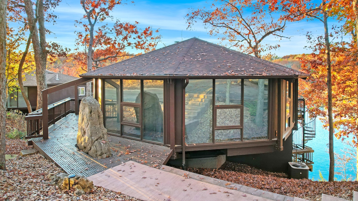 A modern villa is situated by a tranquil lake, featuring large glass walls that allow for natural light and scenic views. Surrounding autumn foliage adds vibrant colors, while a wooden deck extends from the entrance, leading to the lakefront. A natural stone element complements the landscape.