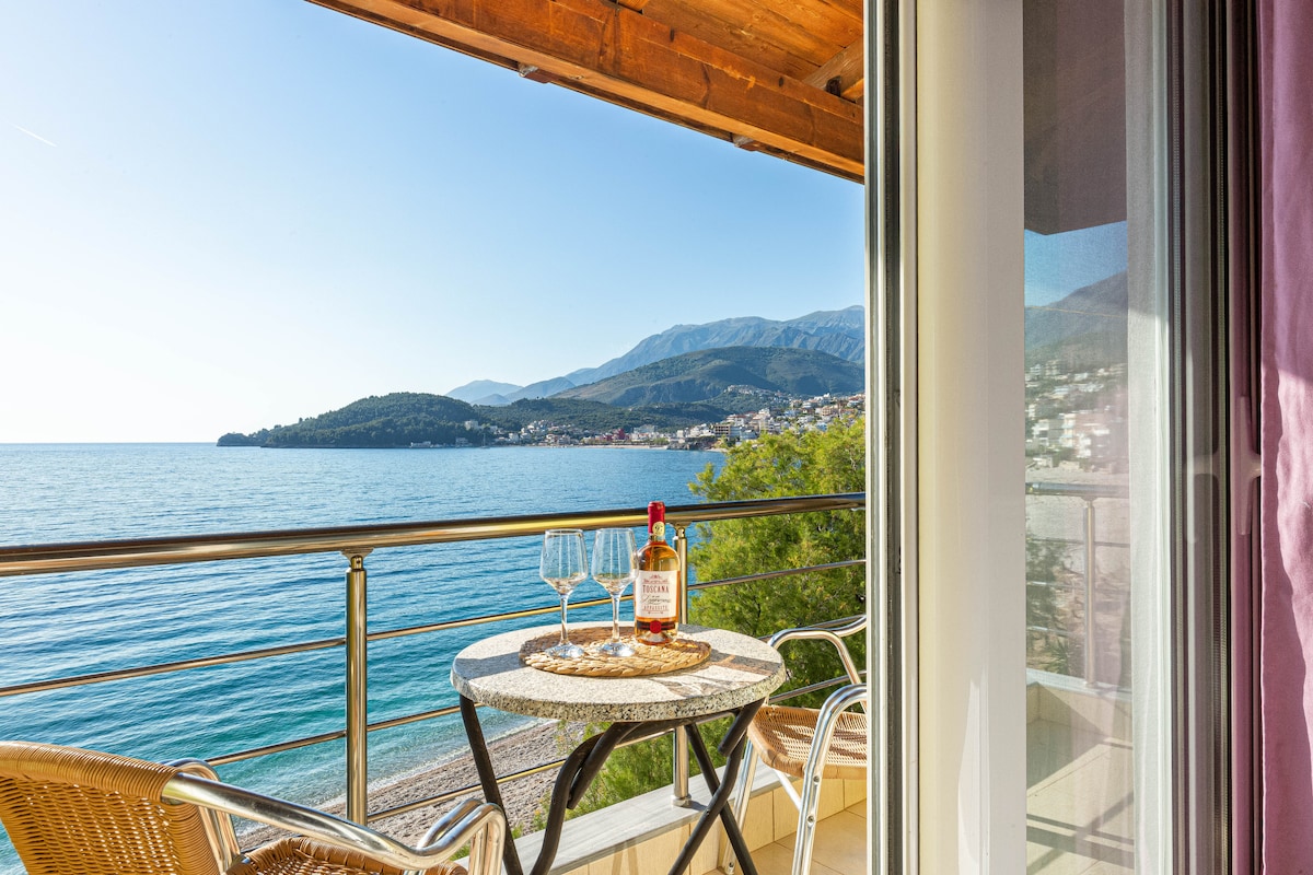 A balcony with a round table set for two holds a bottle of wine and two glasses, overlooking the Ionian Sea. Scenic mountain views are visible in the background, while sunlight reflects off the water, creating a serene atmosphere.
