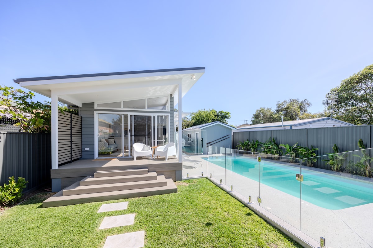 A modern dwelling is presented with a spacious deck that leads to a shared resort-style pool. White chairs are positioned on the deck, surrounded by lush greenery. The pool area features clear glass fencing, allowing for unobstructed views of the inviting water.