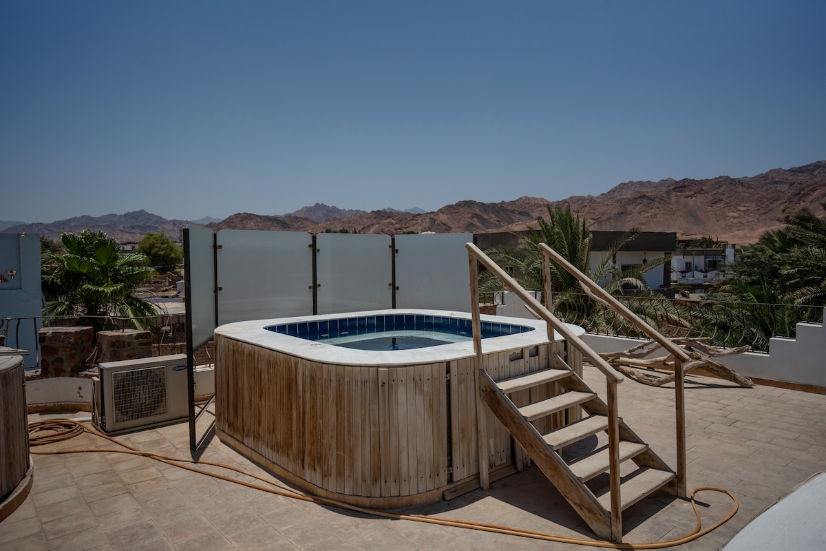 A hot tub is situated on a spacious rooftop terrace, surrounded by wooden decking. Steps lead up to the hot tub area. The backdrop features distant mountains and clear blue skies, creating a serene outdoor environment.