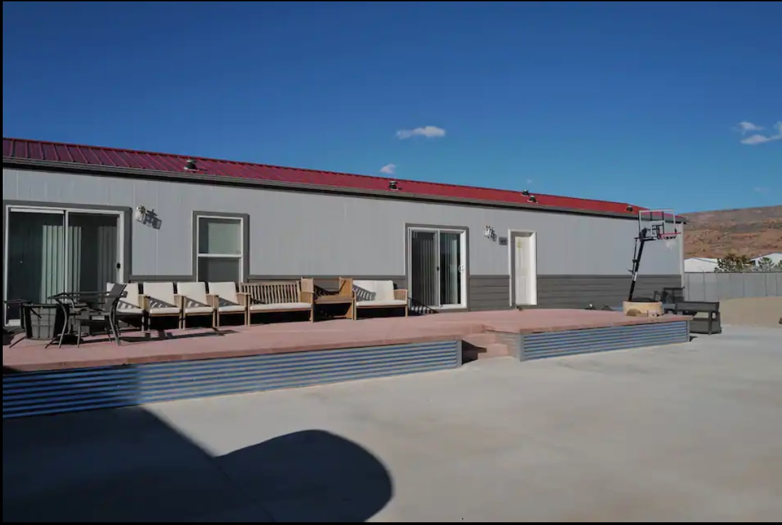 An outdoor area features a spacious deck with various seating options, including lounge chairs and a dining set. A basketball hoop stands nearby on a concrete surface, while large windows provide views of the surrounding landscape.