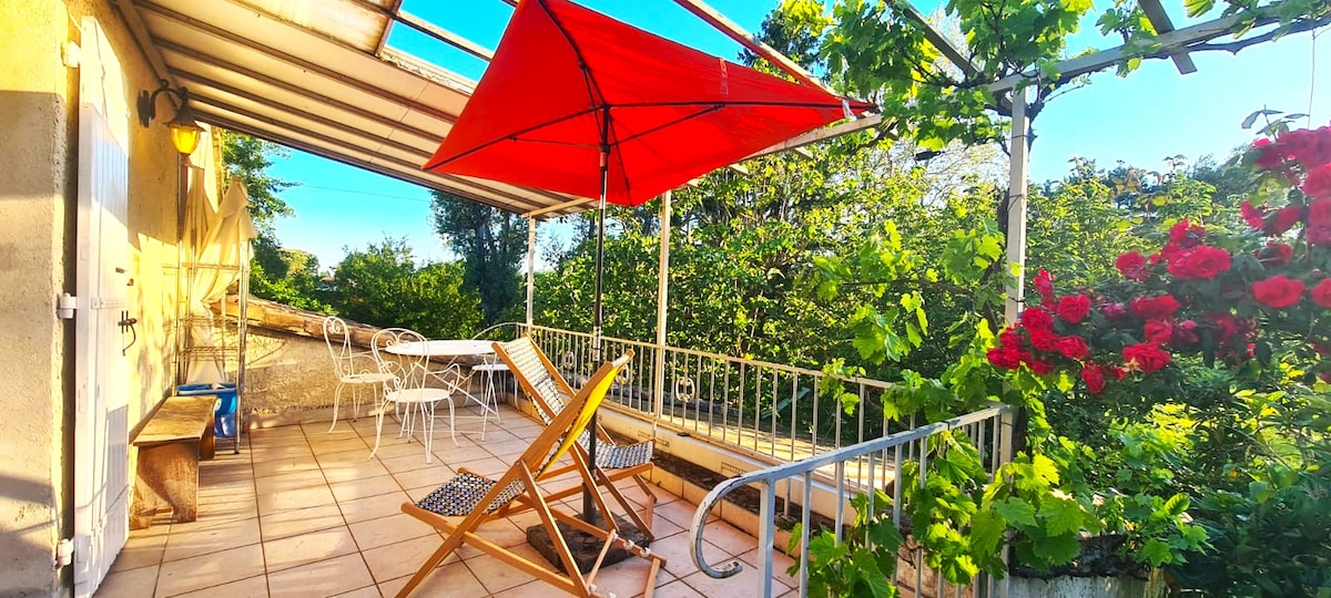 A spacious terrace is seen, featuring a bright red parasol providing shade over a small, round dining table and two white chairs. A wooden lounge chair offers a relaxing spot, surrounded by lush greenery and vibrant flowering plants.