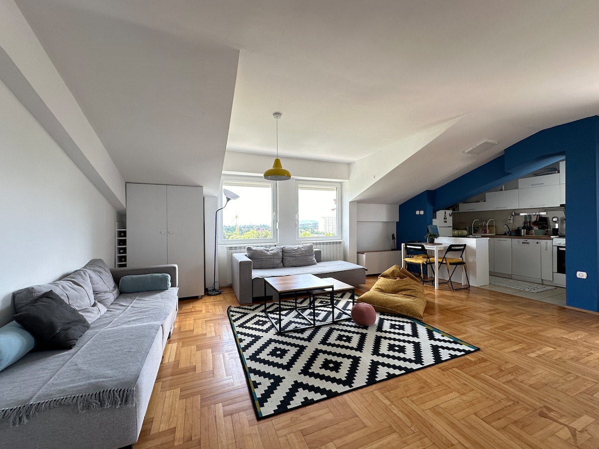 A spacious living area displays a mix of modern and comfortable furnishings. A patterned rug is set on wooden flooring, framing a coffee table surrounded by a sectional sofa and bean bags. Bright light enters through large windows, complementing the open kitchen area.
