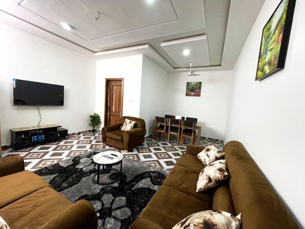 A welcoming living area features two brown sofas adorned with decorative pillows. A flat-screen TV is mounted on the wall opposite a small coffee table. A dining table for four is positioned nearby, complemented by a vibrant piece of artwork on the wall.