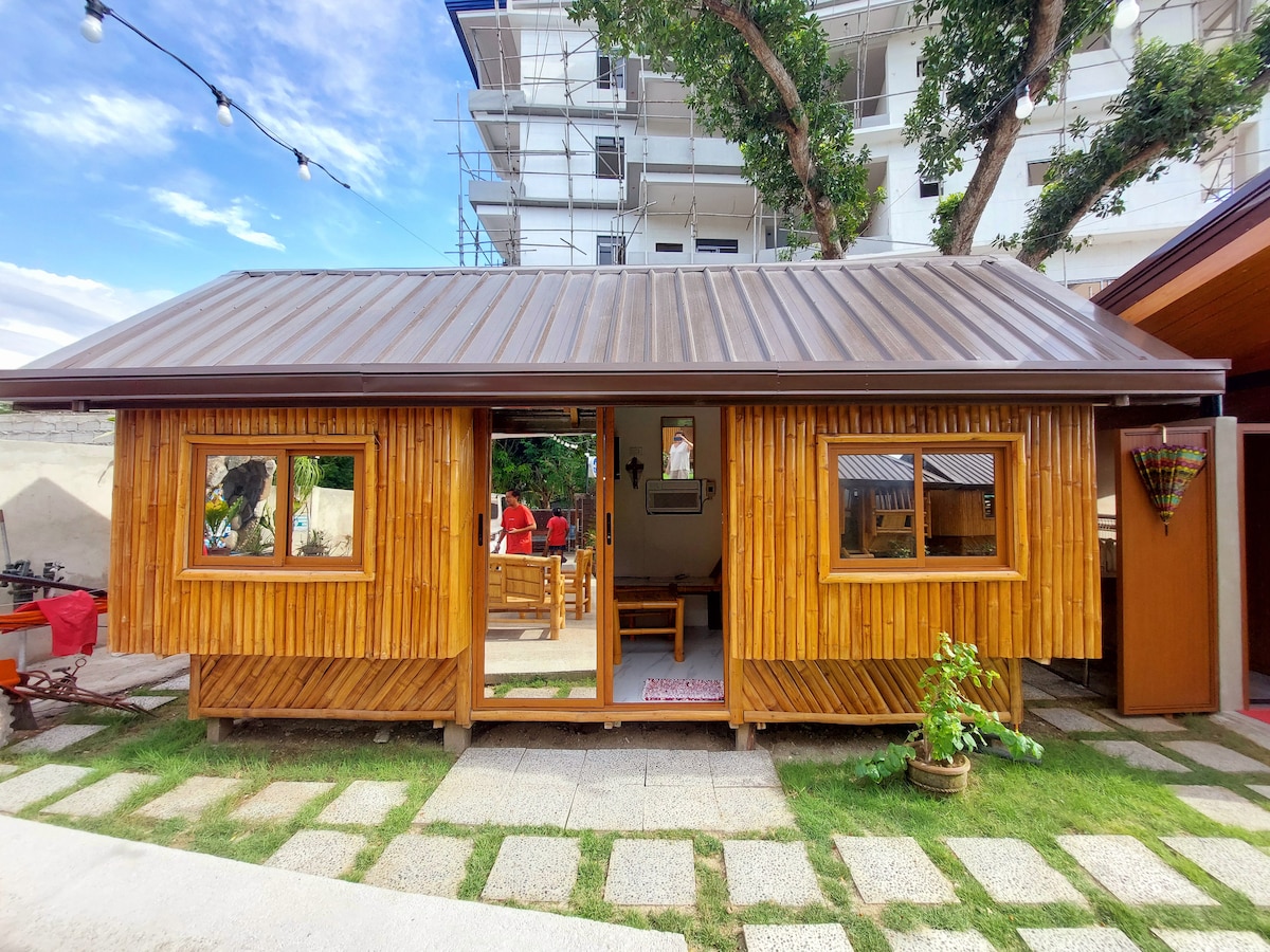 A charming wooden cottage with a sloped metal roof is featured, complemented by large windows that invite natural light. The entrance opens to a cozy outdoor seating area, surrounded by greenery and well-maintained stone pathways.