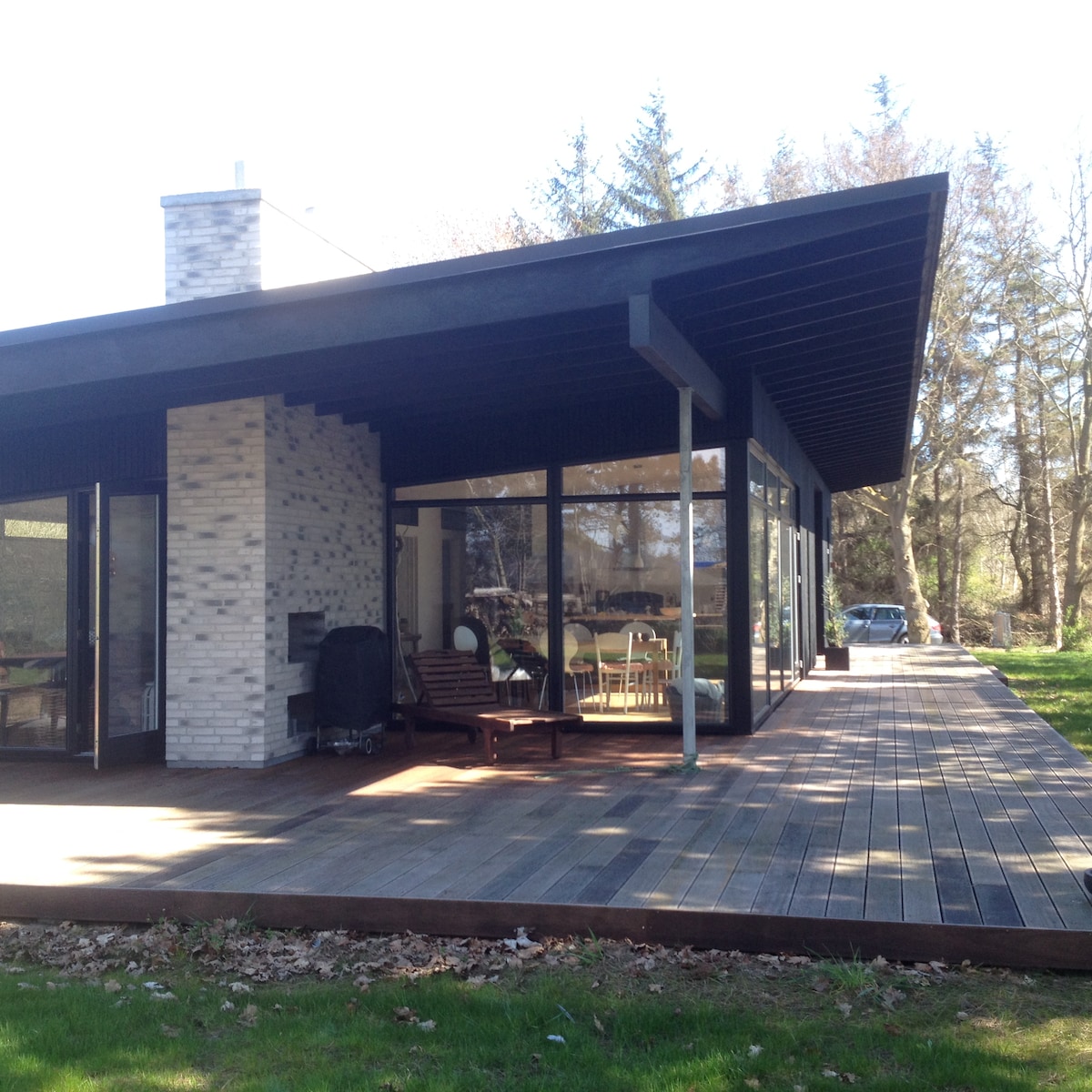 A modern architect-designed house features a spacious deck with a smooth wooden surface. Large glass walls provide ample natural light and showcase a comfortable living space inside. Surrounding greenery adds a peaceful ambiance, enhancing the connection with nature.
