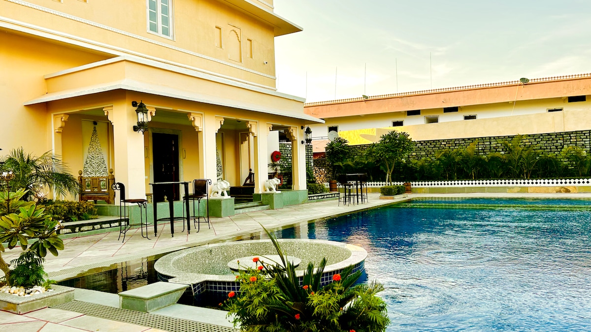An outdoor view of the hotel showcases a large swimming pool surrounded by manicured gardens. Poolside seating is arranged beside the water, and lush greenery is visible throughout the area. The architectural features of the hotel are highlighted against a clear sky.